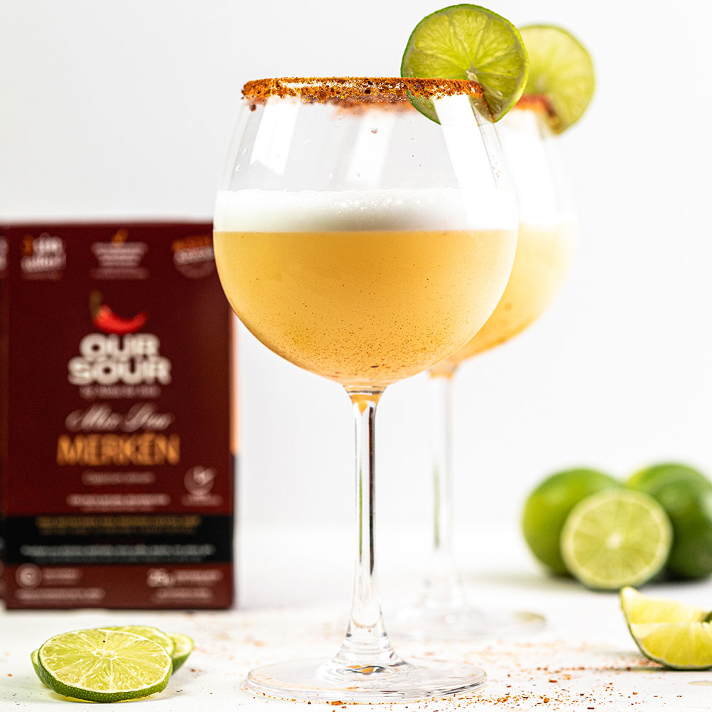 Cóctel sour con merkén servido en copa con borde de ají y rodaja de limón, preparado con Our Sour Merkén. Fondo blanco con estuche del producto desenfocado